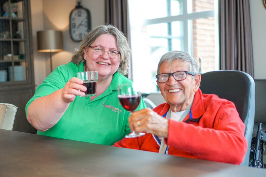 Een bewoner met Alzheimer en een zorgmedewerkster van de kleinschalige particuliere zorglocatie proosten met een glaasje wijn naar de camera