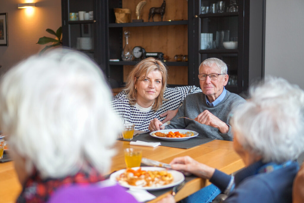 Een locatiemanager en een bewoner met alzheimer kijken vrolijk naar de camera tijdens de lunch die bereid is door de kok