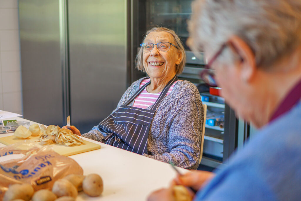 Een bewoonster van de kleinschalige zorglocatie voor mensen met dementie lacht naar de camera tijdens het schillen van de aardappels
