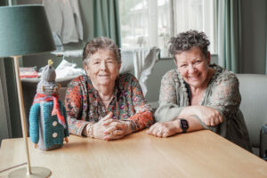 Moeder en dochter zitten samen aan tafel in de huiskamer van Het Warandehuis, de kleinschalige woonzorglocatie voor mensen met dementie van Dagelijks Leven in Bergen op Zoom. Op de tafel staat de Peperbus die moeder zelf gebreid heeft.