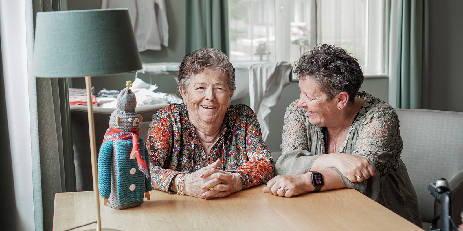 Moeder en dochter zitten samen aan tafel in de huiskamer van Het Warandehuis, de kleinschalige woonzorglocatie voor mensen met dementie van Dagelijks Leven in Bergen op Zoom. Op de tafel staat de Peperbus die moeder zelf gebreid heeft.