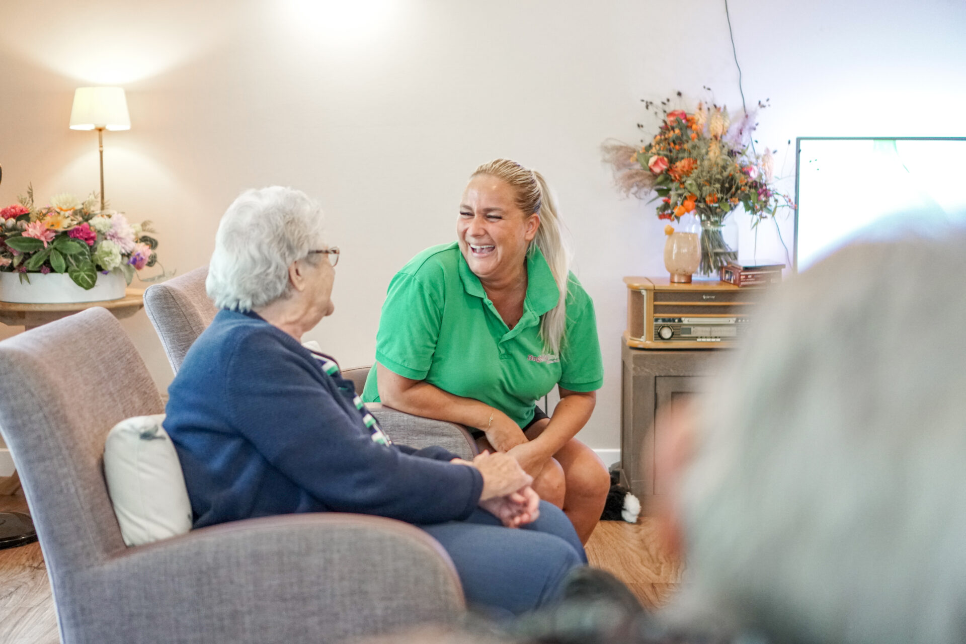 Helpende vacature Venray? Kom werken bij Dagelijks Leven, voor mensen met dementie. Op de foto een helpende met een bewoner, lachend naar elkaar, aan tafel.