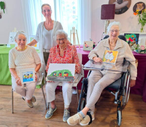 Activiteitenbegeleider met drie deelnemers van de schildersclub die zij in de kleinschalige woonzorglocatie voor mensen met dementie heeft opgezet. Ze tonen trots hun schilderijen.