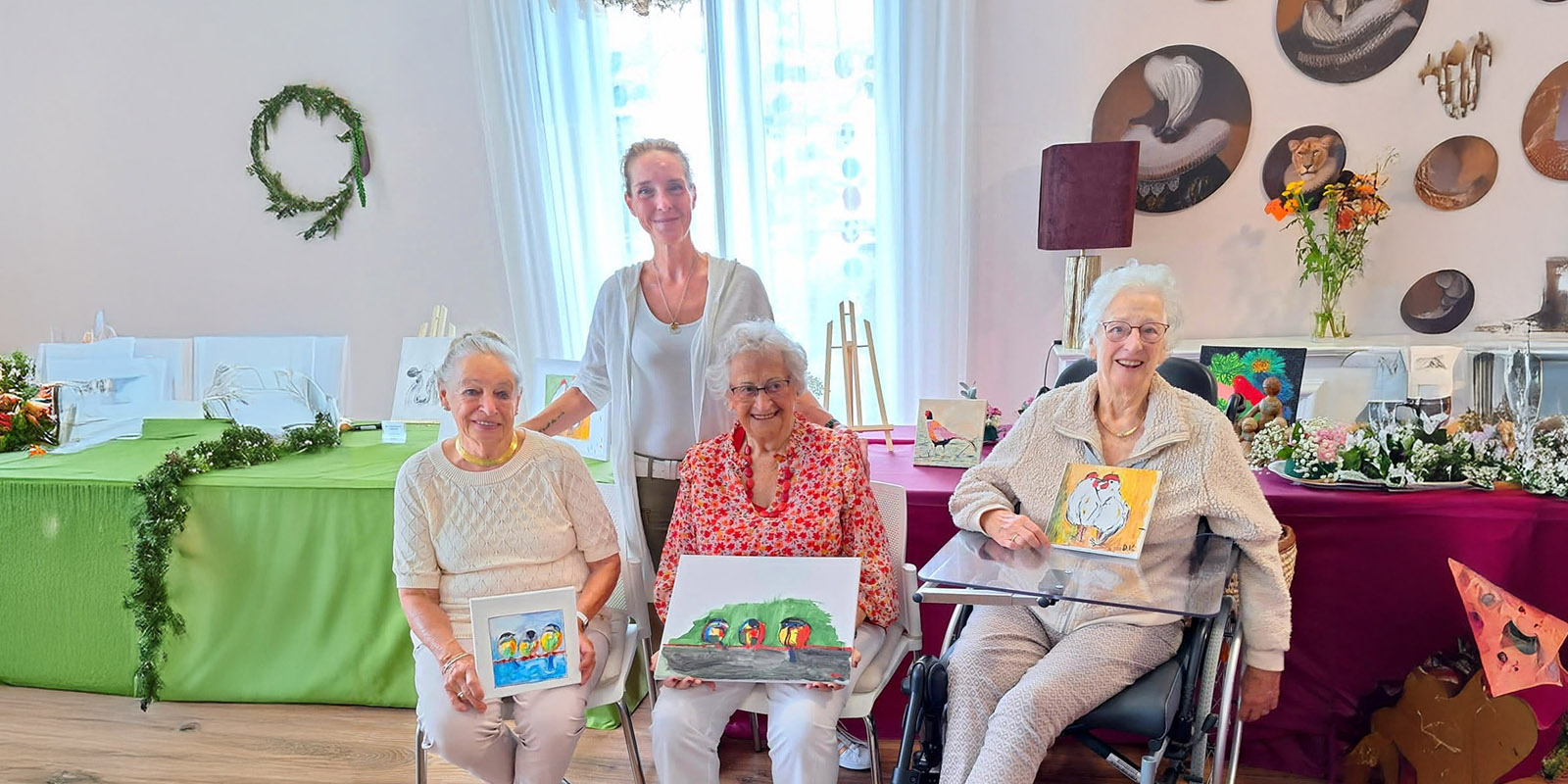 Activiteitenbegeleider met drie deelnemers van de schildersclub die zij in de kleinschalige woonzorglocatie voor mensen met dementie heeft opgezet. Ze tonen trots hun schilderijen.