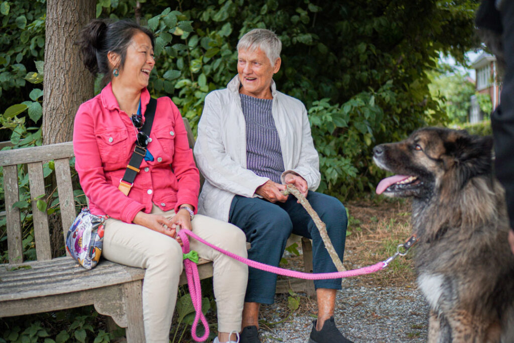 Een vrijwilligster en een bewoonster van de woonzorglocatie van Dagelijks Leven voor mensen met dementie laten samen de hond uit en zitten op het bankje te rusten.