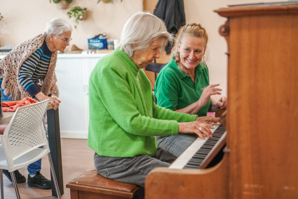 De bewoonster en activiteitenbegeleider van de woonzorglocatie van Dagelijks Leven voor mensen met dementie spelen samen piano in de activiteitenruimte