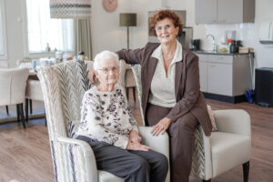 Foto van dochter met moeder, die in Het RIngbaanhuis woont, onze kleinschalige woonzorglocatie voor mensen met dementie in Tilburg. Moeder zit in fauteuil, dochter op de stoelleuning. 